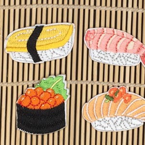 May include: Four embroidered sushi patches with different fillings: a yellow omelet, a pink shrimp, orange fish roe, and a salmon nigiri. The patches are on a bamboo mat background.