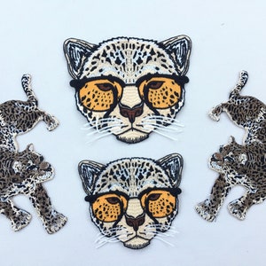 May include: Set of five embroidered iron-on patches featuring a cheetah wearing sunglasses. Two patches are of the cheetah's face, and three patches are of the cheetah's full body.