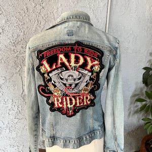 Pode incluir: Casaco de ganga azul claro com um grande patch bordado nas costas. O patch apresenta as palavras "Freedom to Ride Lady Rider" com um gr&aacute;fico de motor de motocicleta e detalhes florais.