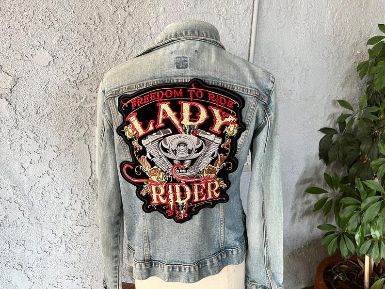 Pode incluir: Casaco de ganga azul claro com um grande patch bordado nas costas. O patch apresenta as palavras "Freedom to Ride Lady Rider" com um gr&aacute;fico de motor de motocicleta e detalhes florais.