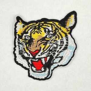 May include: Embroidered iron-on patch of a tiger's face with yellow, black, white, and red details. The tiger has a fierce expression with its mouth open and teeth showing.