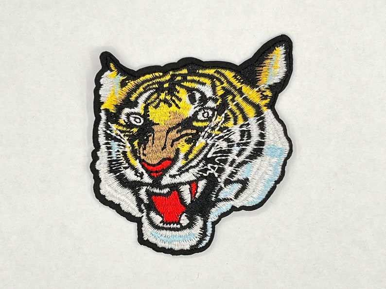 May include: Embroidered iron-on patch of a tiger's face with yellow, black, white, and red details. The tiger has a fierce expression with its mouth open and teeth showing.