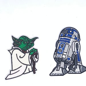 May include: Two embroidered iron-on patches featuring Star Wars characters. The first patch is of Yoda, a green alien with large ears, wearing a white robe and holding a brown walking stick. The second patch is of R2-D2, a blue and white robot with a dome-shaped head.