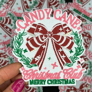 May include: White embroidered patch with a red bow, green leaves, and the text "Candy Cane Christmas Club Merry Christmas".