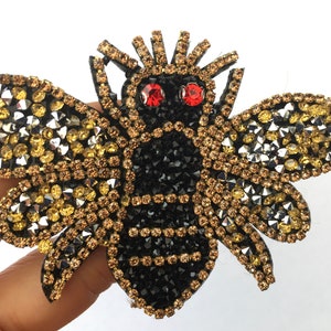 May include: A black and gold bee-shaped patch with rhinestones. The bee has red eyes and is surrounded by gold and silver rhinestones.