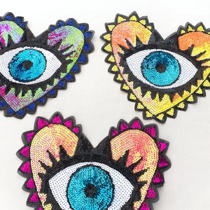 May include: Three heart-shaped patches with a large eye design. The eyes are made of sequins and have a white iris and a blue pupil. The hearts are made of iridescent sequins in shades of pink, yellow, and blue.