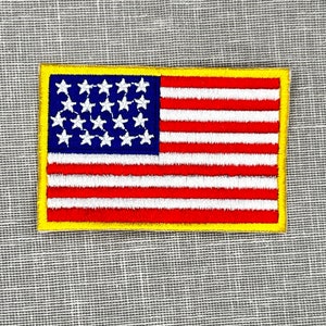 May include: Embroidered rectangular patch featuring the American flag. The flag design includes a blue canton with white stars, red and white stripes, and a yellow border. The patch is likely for clothing or accessories.