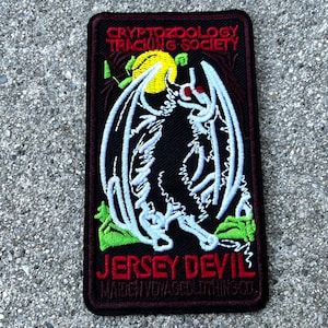May include: A black embroidered patch with a white outline of a winged creature with red eyes. The patch has the text "CRYPTOZOOLOGY TRACKING SOCIETY" at the top and "JERSEY DEVIL" at the bottom. The text "MATCH YOUR STYLE WITH THIS SO!" is in smaller font below the main text.