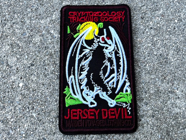May include: A black embroidered patch with a white outline of a winged creature with red eyes. The patch has the text "CRYPTOZOOLOGY TRACKING SOCIETY" at the top and "JERSEY DEVIL" at the bottom. The text "MATCH YOUR STYLE WITH THIS SO!" is in smaller font below the main text.