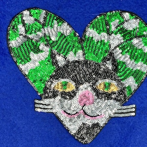May include: A black and white cat with green and silver sequins, shaped like a heart. The cat has large green eyes and a pink nose.