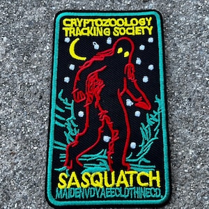 May include: Black embroidered patch with a green border. It features a red and black sasquatch walking in a snowy forest. The text "CRYPTOZOOLOGY TRACKING SOCIETY" is at the top and "SASQUATCH MAIDENVDYAEECLOTHINECO" is at the bottom.