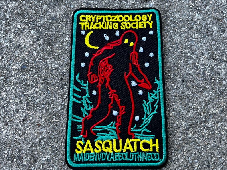 May include: Black embroidered patch with a green border. It features a red and black sasquatch walking in a snowy forest. The text "CRYPTOZOOLOGY TRACKING SOCIETY" is at the top and "SASQUATCH MAIDENVDYAEECLOTHINECO" is at the bottom.