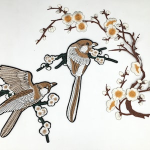 May include: Two embroidered patches featuring brown birds perched on branches with white blossoms. The birds have brown and white feathers and are surrounded by delicate white flowers.