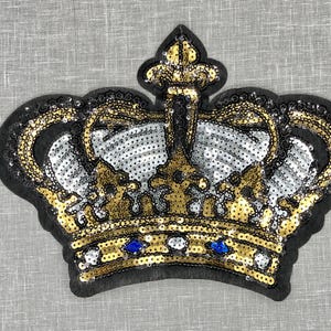 May include: A gold and silver sequined crown patch with blue jewels. The crown is on a gray fabric background.
