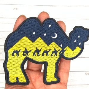 May include: A yellow and blue embroidered patch of a camel with a starry night sky and mountains in the background. The camel is carrying a line of smaller camels across the desert.