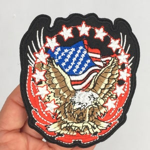 May include: Embroidered patch featuring a bald eagle with outstretched wings holding an American flag. The eagle is surrounded by a circle of white stars on a black background.