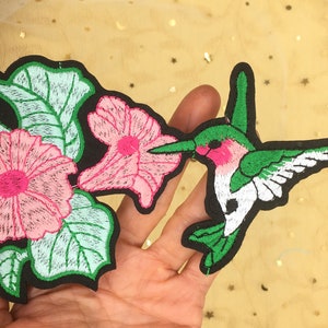 May include: Embroidered iron-on patch featuring a hummingbird with green and white wings, a pink body, and a long beak, hovering near pink flowers with green leaves.