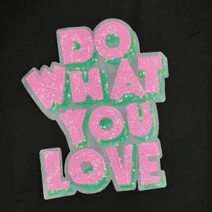 May include: A pink sequined patch with the words "DO WHAT YOU LOVE" in large, bubble letters. The letters are outlined in green and set against a sheer, white background. The patch is on a black background.
