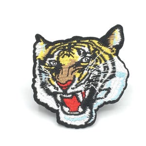 May include: Embroidered iron-on patch featuring a tiger's face with a fierce expression. The tiger has black, white, and yellow stripes and a red open mouth.