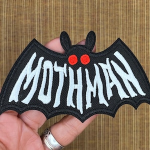 May include: Black embroidered patch in the shape of a bat with the word "MOTHMAN" in white, stylized lettering. The bat has red eyes and small ears. The patch is held in a hand.