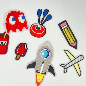 May include: A set of 8 iron-on patches featuring various designs, including a red Pac-Man, a red and white target with three blue arrows, a red and white popsicle, a red and white can with the word "COLA" on it, a gray rocket with a blue window and yellow flames, a yellow pencil with a pink eraser, a gray and white airplane, and a red and white pencil.
