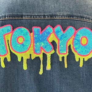May include: Denim jacket with a sequined "TOKYO" design on the back. The letters are turquoise and blue with a pink outline, and a neon yellow dripping effect. The jacket is made of blue denim.