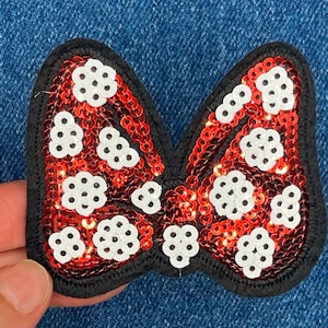 May include: A sequined bow-shaped patch with a black border. The bow is filled with red sequins and white floral-shaped accents. The patch is likely for clothing or accessories.