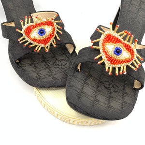 May include: A pair of black fabric sandals with a beaded heart-shaped evil eye design. The sandals have a simple strap design and are adorned with red, blue, and gold beads.