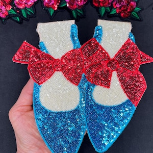 May include: A pair of blue and white sequined shoes with a red bow on top. The shoes are decorated with sequins and have a sparkly finish.