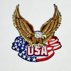 May include: Embroidered patch featuring a majestic eagle with outstretched wings. The eagle is brown and white, clutching a banner with "USA" in red, white, and blue. The background includes the American flag.