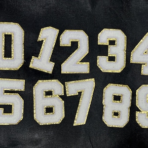 May include: White felt numbers with gold glitter trim, from 0 to 9, on a black background.