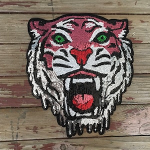 May include: A sequined tiger face patch with a black border. The tiger has a red and pink face, green eyes, and white and black stripes. The mouth is open, revealing red sequins.