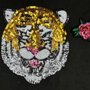 May include: A gold and silver sequined tiger face applique with a pink tongue and black eyes. The tiger is surrounded by a black background.