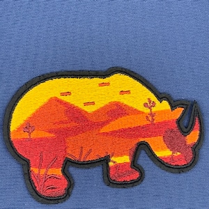 May include: A black and orange embroidered patch of a rhinoceros silhouette with a desert landscape inside. The landscape features a yellow sky with orange mountains and red sand.