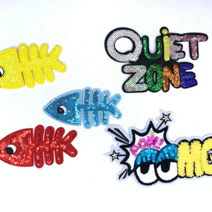 May include: Five colorful iron-on patches with sequin details. The patches include a blue fishbone, a yellow fishbone, a red fishbone, a colorful "Quiet Zone" patch, and a yellow and blue "OMG!" patch with large eyes.