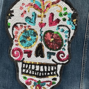 May include: A denim jacket back with a large, sequined sugar skull patch. The skull features a white base with colorful sequins in teal, pink, red, and gold, and a black outline. The jacket is a medium wash denim.