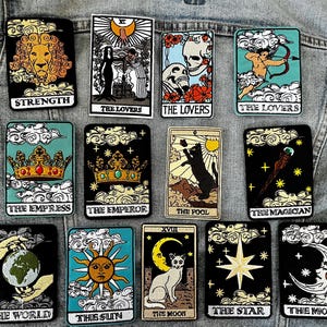 May include: A set of 14 tarot card patches featuring various tarot card imagery, including the Strength, Lovers, Empress, Emperor, Fool, Magician, World, Sun, Moon, Star, and Magician cards. The patches are made of felt and have a colorful, whimsical design.