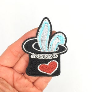 May include: A black felt embroidered patch of a magic hat with a white and blue rabbit sticking out of it. A red heart is on the front of the hat.