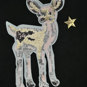 May include: A light pink and gold sequined deer applique with a gold star. The deer is standing on all four legs and has a long neck. The star is to the right of the deer.