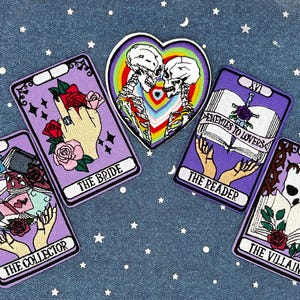 May include: Five embroidered iron-on patches featuring tarot card imagery. The patches are purple with white and black embroidery. The cards depicted are The Collector, The Bride, The Reader, Enemies to Lovers, and The Villain.