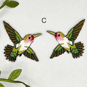 May include: Two green, white, and pink embroidered hummingbird patches. The hummingbirds are facing opposite directions.