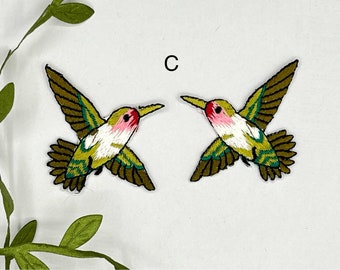 May include: Two green, white, and pink embroidered hummingbird patches. The hummingbirds are facing opposite directions.