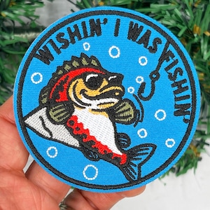 May include: Round blue patch with the text "Wishin' I Was Fishin'". The patch features a cartoon fish with red, yellow, and white details, a fishing hook, and bubbles. The patch is held in a hand.
