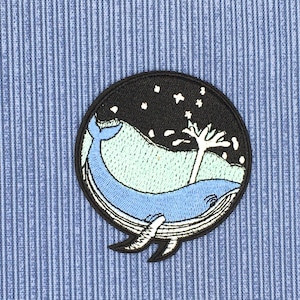 May include: Embroidered patch featuring a blue whale swimming in a circle with a starry night sky and a white tree in the background.