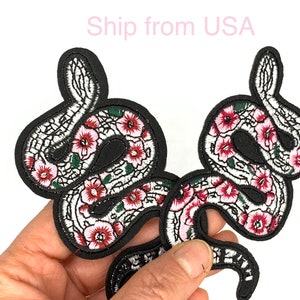 May include: Two black embroidered iron-on patches of snakes with pink and green flowers. The snakes are intertwined and have a detailed design.