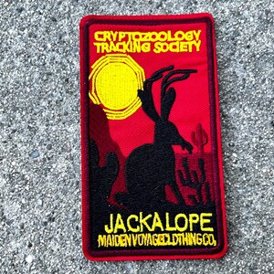 May include: A red embroidered patch with a black silhouette of a jackalope with antlers, a yellow sun, and the text "Cryptozoology Tracking Society" and "Jackalope Maiden Voyage Clothing Co."