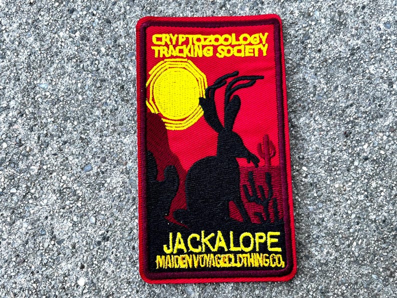 May include: A red embroidered patch with a black silhouette of a jackalope with antlers, a yellow sun, and the text "Cryptozoology Tracking Society" and "Jackalope Maiden Voyage Clothing Co."