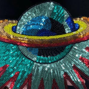 May include: A colorful sequined applique with a celestial design. The applique features a rainbow of colors, including red, yellow, blue, and green, arranged in a circular pattern. The design is embellished with black thread.