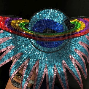 May include: A sequined applique in the shape of a stylized planet with a rainbow-colored ring. The planet is primarily blue, with a central dark blue area. The wings are turquoise with pink accents. The applique is on a black background.