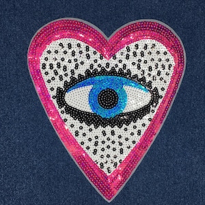 May include: A pink heart-shaped patch with a blue evil eye design. The heart is covered in sequins and the eye is surrounded by black sequins.